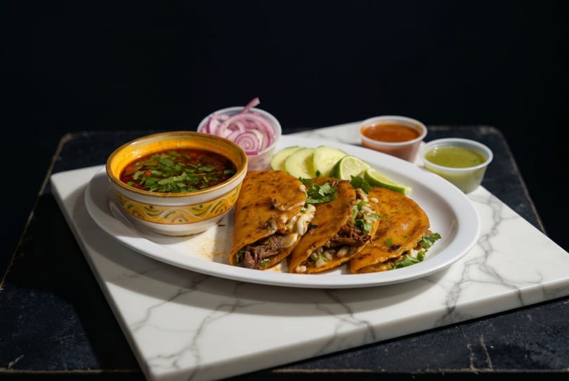 Birria tacos with consommé, red onions, cucumbers, limes, and two sauces on a white plate.