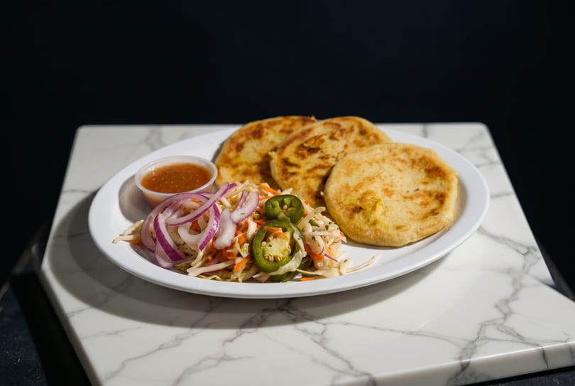 Three golden pupusas with curtido, red onions, jalapeños, and a side of salsa on a white plate.