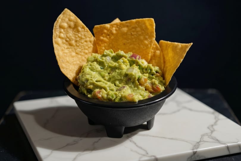 A black bowl of chunky guacamole with tortilla chips on a white marble slab.